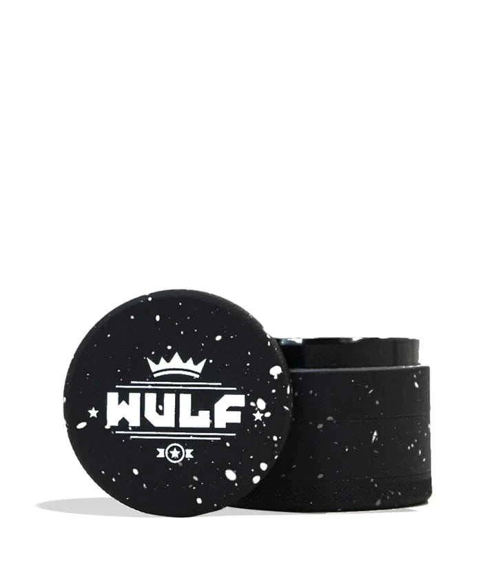 Front angled view of the Wulf 4-Piece 65mm Aerospace Aluminum Grinder in black with white splatter. Crafted from durable aerospace-grade metal, this large multi-chamber grinder effortlessly shreds dry botanicals to the perfect consistency for rolling or packing.