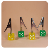 Front angled view of the Dice Roach Clip 10 Piece Pack in yellow and green. Featuring a sturdy metal alligator clip topped with a classic casino-style die, these helpful smoking accessories securely hold joints to prevent burned fingers.