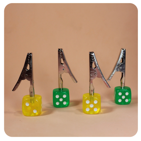Front angled view of the Dice Roach Clip 10 Piece Pack in yellow and green. Featuring a sturdy metal alligator clip topped with a classic casino-style die, these helpful smoking accessories securely hold joints to prevent burned fingers.