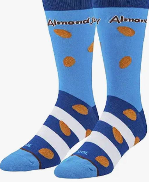 Front view of a pair of Crazy Socks featuring officially licensed Almond Joy graphics. These vibrant novelty crew socks showcase the classic white and red candy logo alongside detailed almond and coconut graphics against a rich royal blue background.
