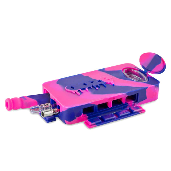 Front angled view of the Ooze Slugger Dugout Travel Kit in pink and purple. This pocket-sized silicone travel piece neatly contains all your necessary concentrate accessories inside an impact-resistant, discreetly flat block.