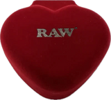 Angled overhead view of the closed red velvet heart-shaped jewelry box featuring an embossed gold RAW logo on the lid. This luxury protective case keeps your delicate borosilicate glass smoke stone safe and pristine between desktop sessions.