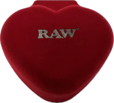 Angled overhead view of the closed red velvet heart-shaped jewelry box featuring an embossed gold RAW logo on the lid. This luxury protective case keeps your delicate borosilicate glass smoke stone safe and pristine between desktop sessions.