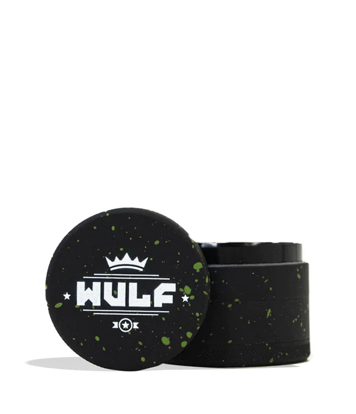 Front angled view of the Wulf 4-Piece 65mm Aerospace Aluminum Grinder in black with green splatter. Crafted from durable aerospace-grade metal, this large multi-chamber grinder effortlessly shreds dry botanicals to the perfect consistency for rolling or packing.